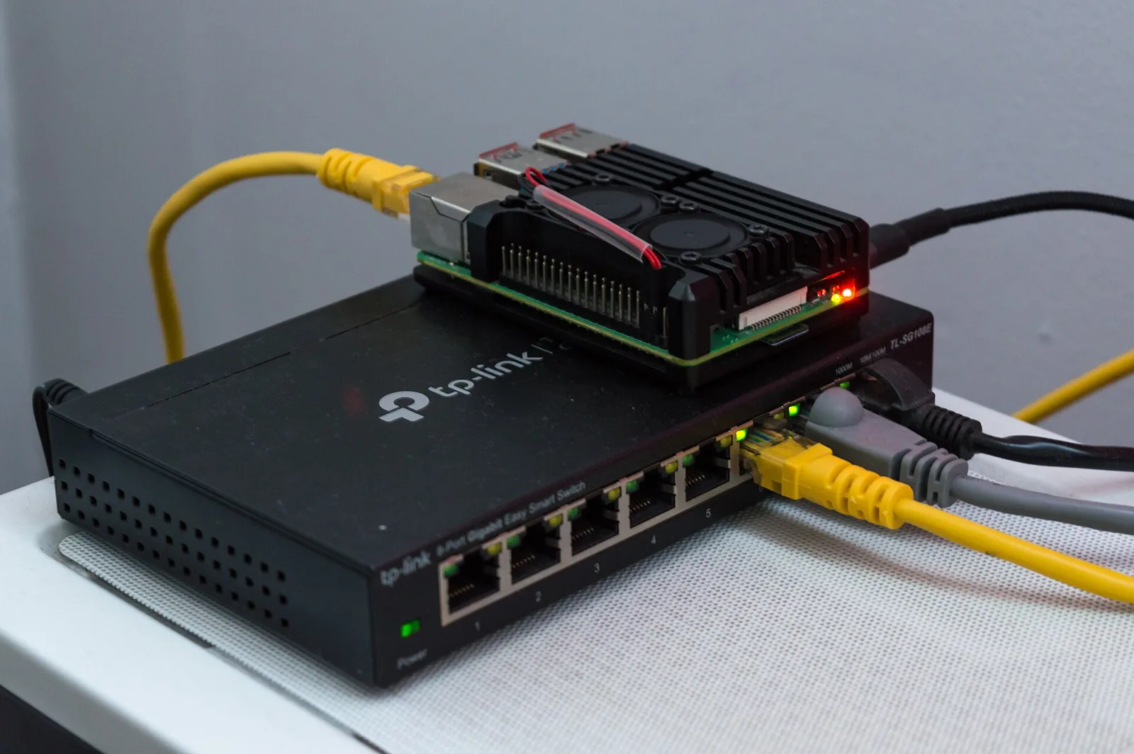 Raspberry Pi in a dark case sitting on a small network switch, yellow Ethernet cables plugged in